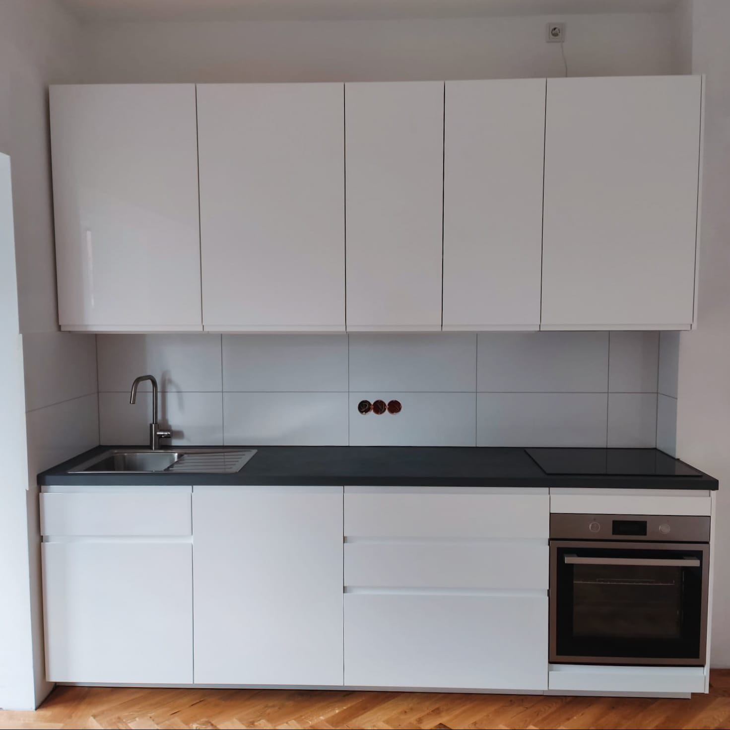 Biała, minimalistyczna zabudowa kuchenna z ciemnym blatem i płytkami w tle. Widoczny zlew, płyta indukcyjna i piekarnik. Parkiet na podłodze.