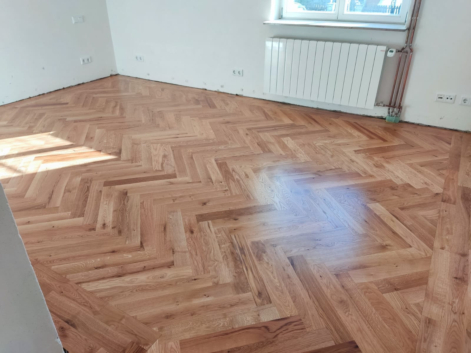 Ukończona podłoga z parkietu ułożonego w jodełkę, widoczna w jasnym, pustym pokoju z grzejnikiem i gniazdkami elektrycznymi na ścianie.