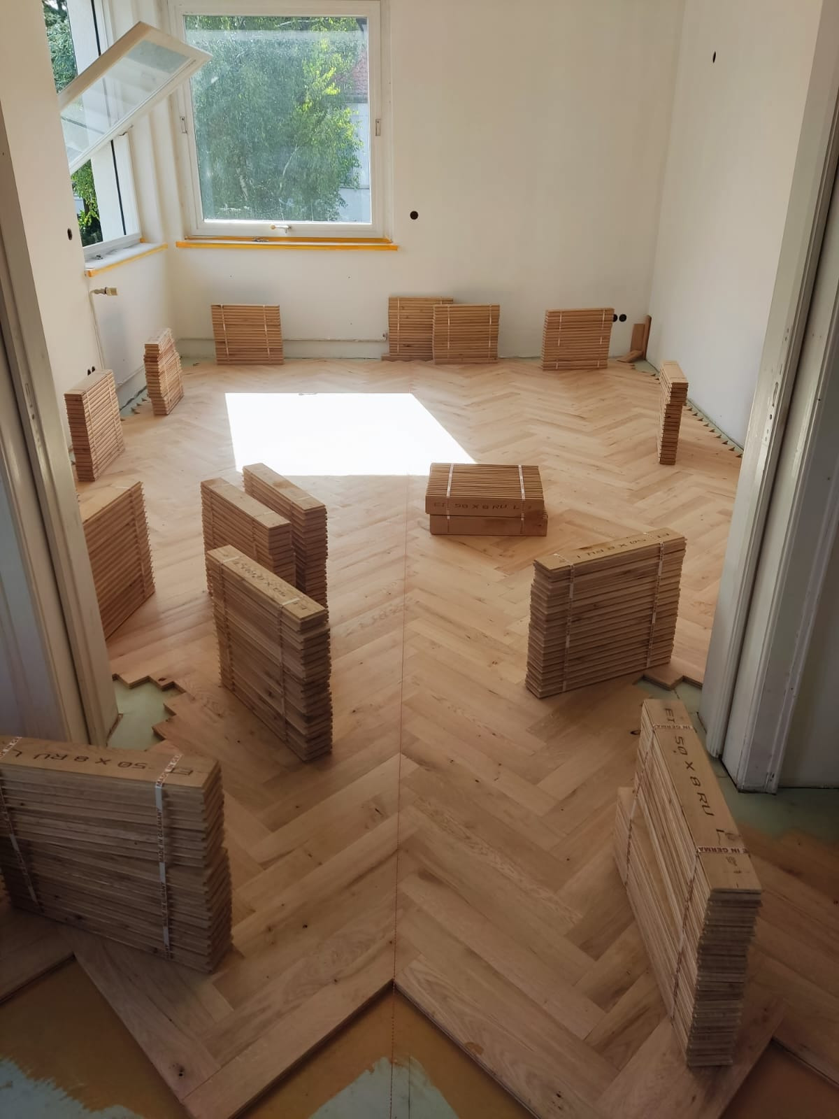 Układanie parkietu w jodełkę w jasnym pokoju z otwartym oknem. Pakiety desek ułożone na podłodze, częściowo pokrytej parkietem. Widoczne niedokończone fragmenty podłogi.