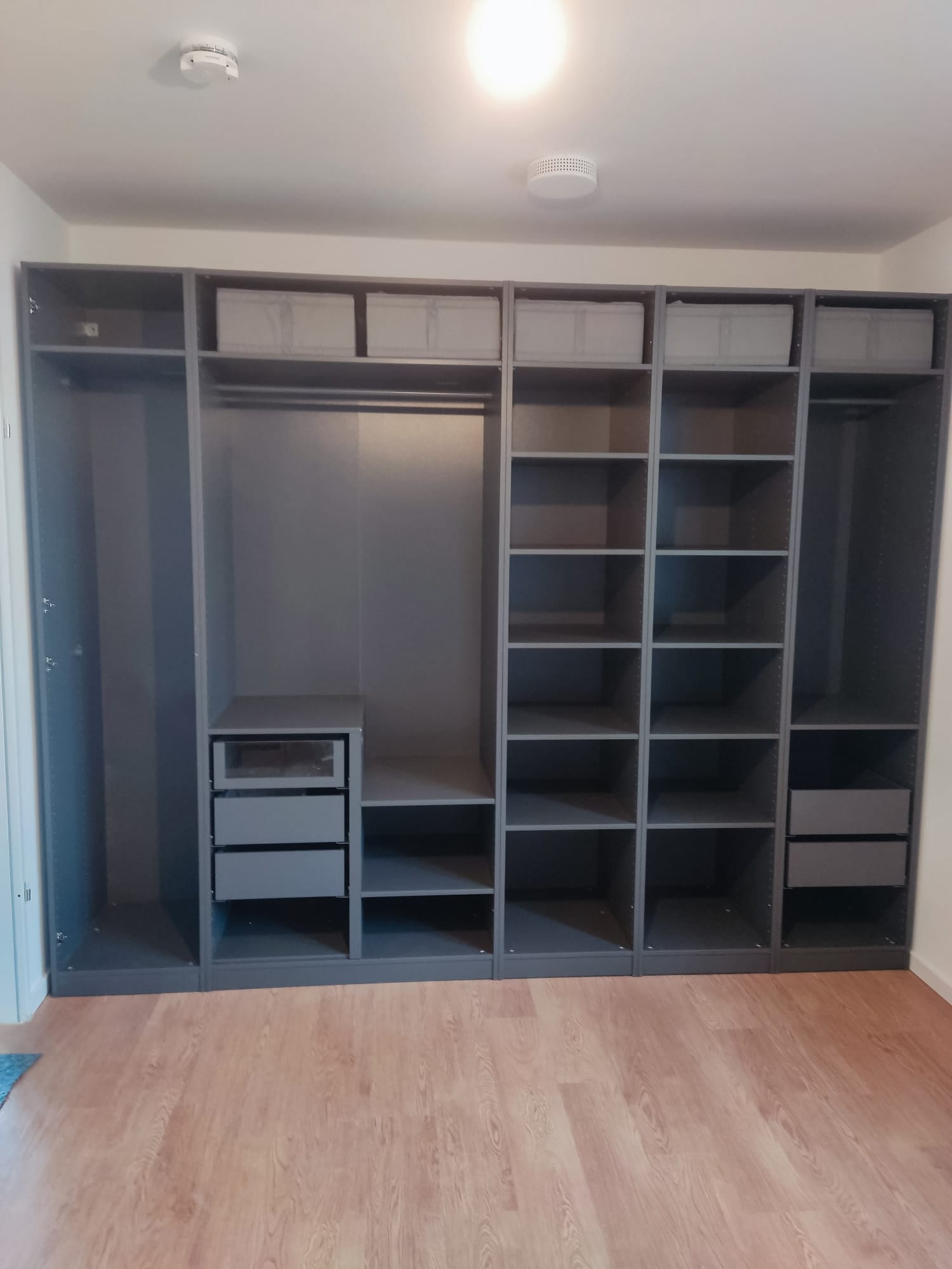 Szara, modułowa szafa wnękowa z półkami, szufladami i miejscem na wieszaki, uzupełniona tekstylnymi pojemnikami na górnych półkach, na tle paneli podłogowych.