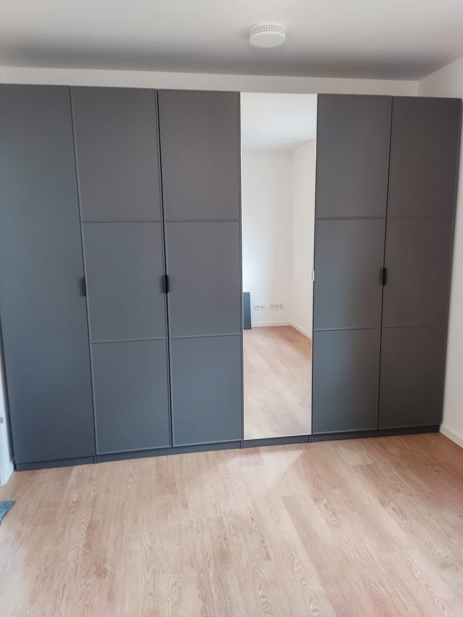 Szara szafa z lustrem wkomponowana w ścianę, widok na drewnianą podłogę i fragment wnętrza pokoju. Minimalistyczny design, proste uchwyty.