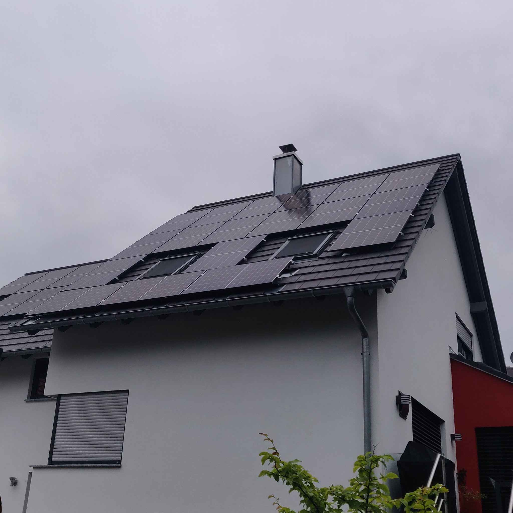 Instalacja paneli fotowoltaicznych na dachu dwuspadowym z oknami dachowymi, widok z dołu na szarym tle nieba.