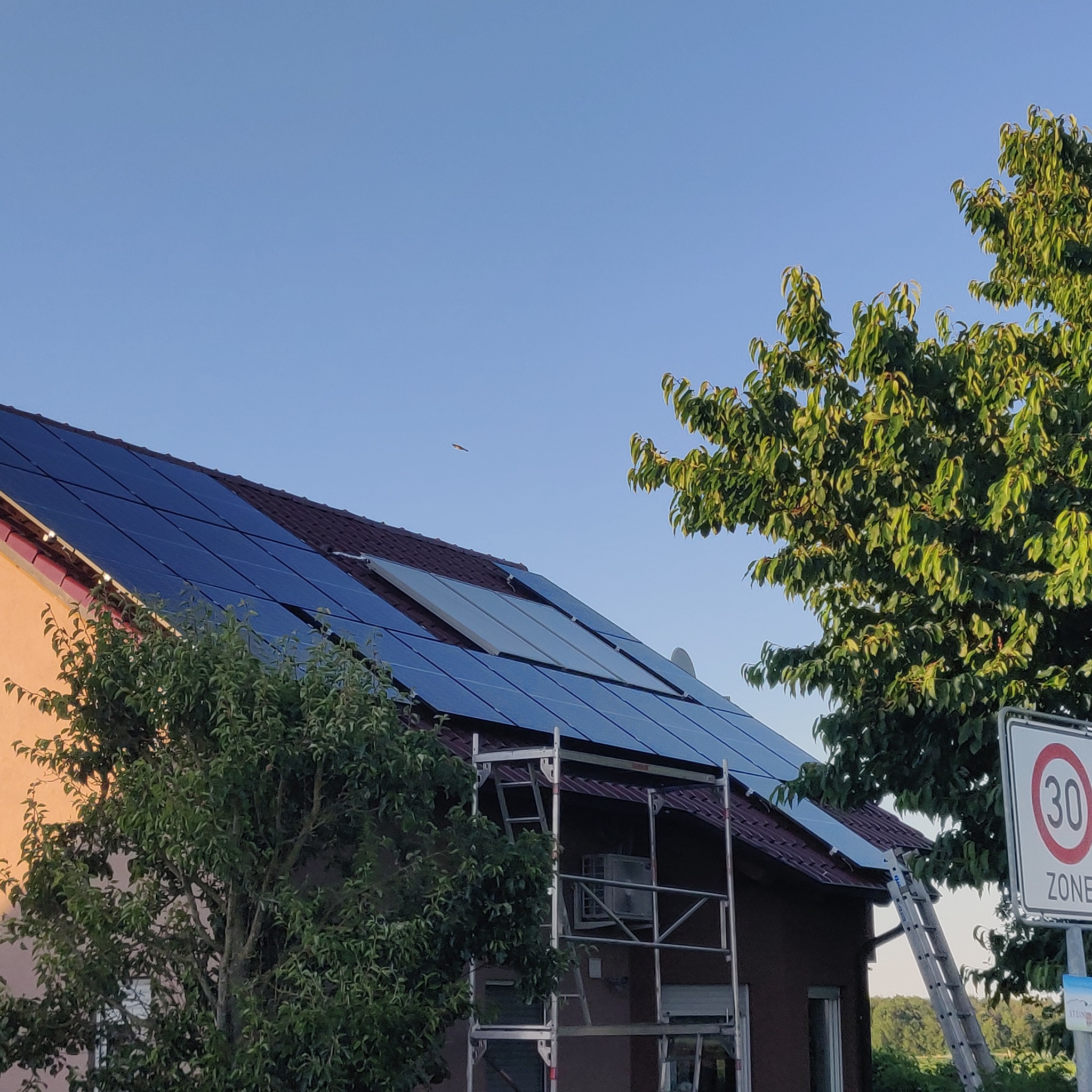 Instalacja paneli słonecznych na dachu domu jednorodzinnego, widoczne rusztowanie i drabina, znak ograniczenia prędkości w tle, częściowo zamontowane panele.