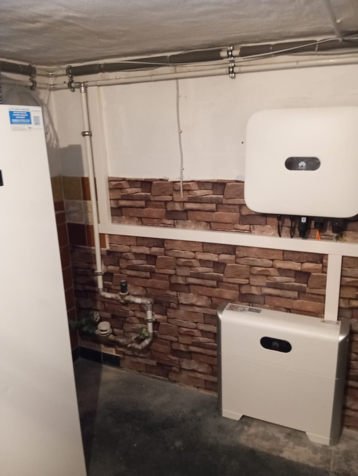 Biały inwerter i akumulator w pomieszczeniu z imitacją ceglanej ściany, widoczne rury instalacyjne pod sufitem.