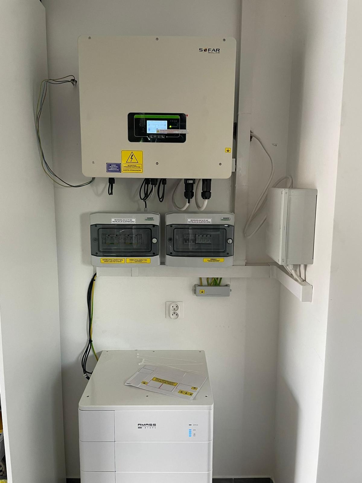 Zamontowany inwerter SOFAR, dwie skrzynki rozdzielcze PV AC, biały magazyn energii AMASS z instrukcjami na górze, zainstalowane na białej ścianie.