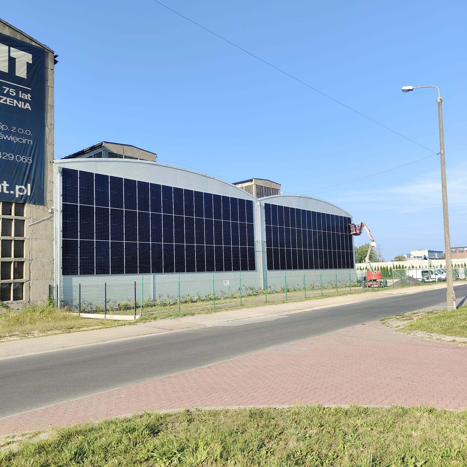 Duży budynek przemysłowy z zamontowanymi panelami słonecznymi na elewacji, widoczny podnośnik koszowy podczas prac instalacyjnych, ujęcie z poziomu ulicy w słoneczny dzień.
