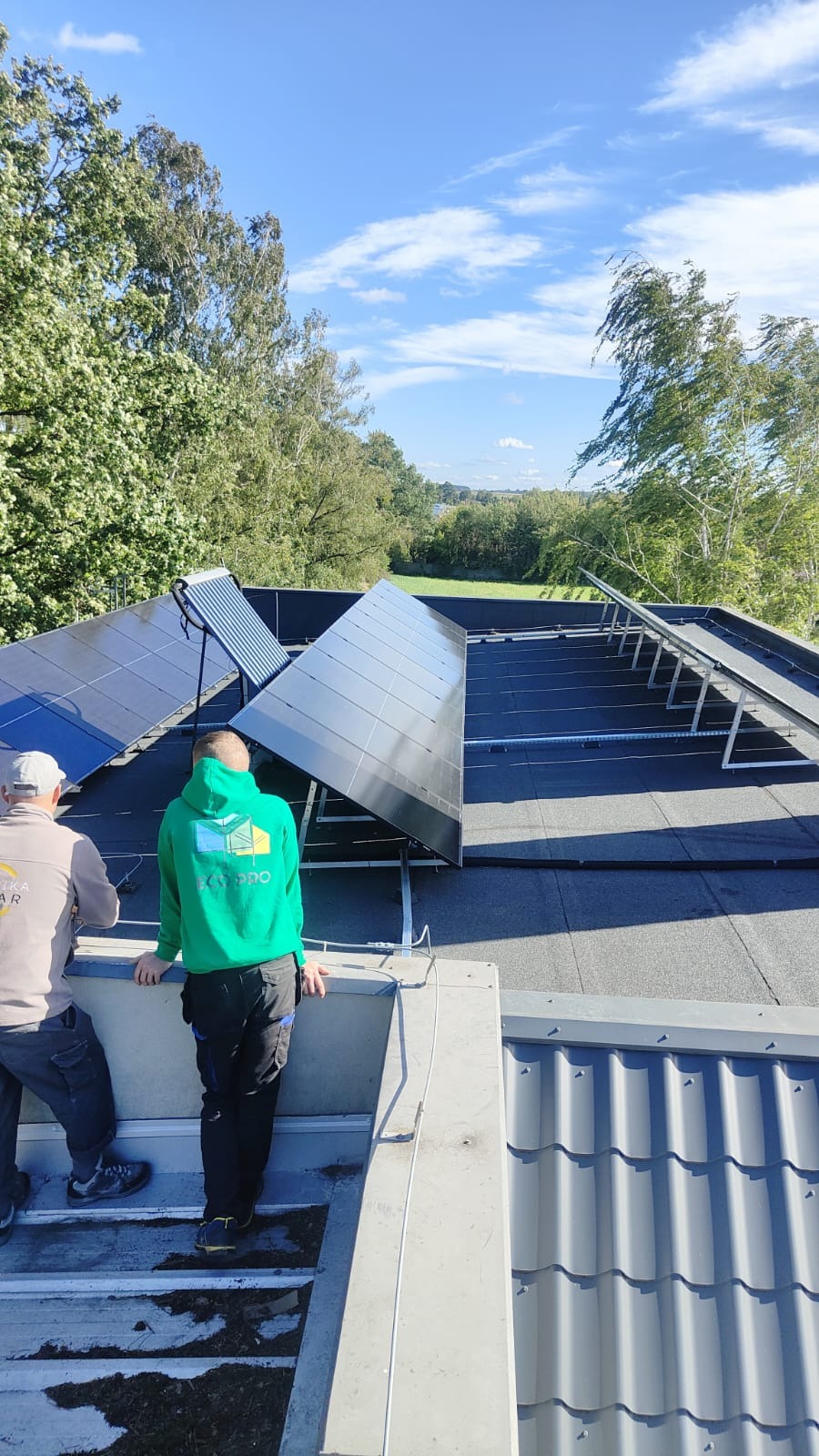 Montaż paneli słonecznych na płaskim dachu pokrytym papą, widoczni dwaj monterzy w odzieży roboczej, jeden w zielonej bluzie firmowej, drugi w szarej, w tle drzewa i błękitne niebo z chmurami.