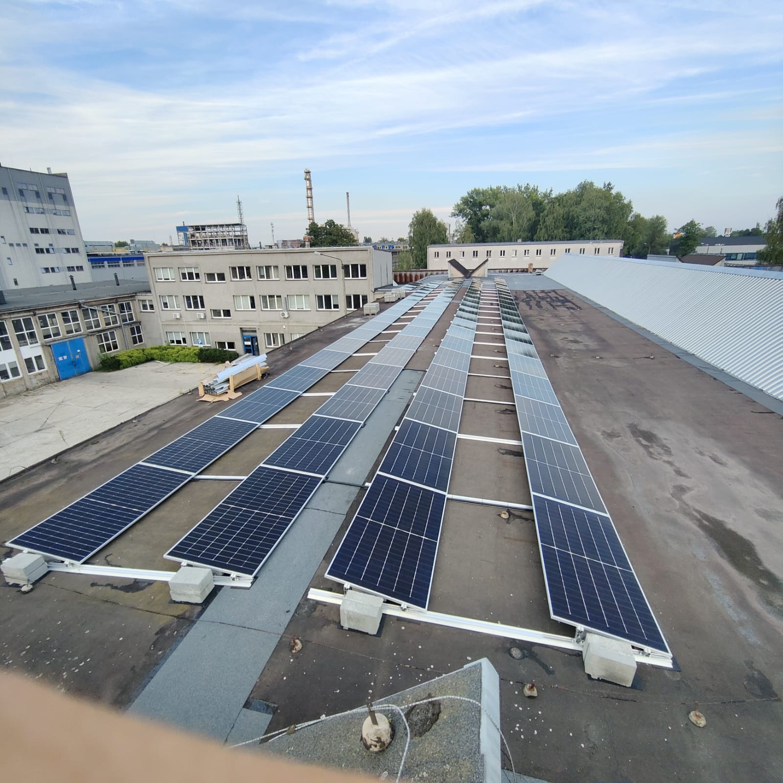 Rząd paneli słonecznych na płaskim dachu budynku przemysłowego, widok z góry, z widocznymi elementami montażowymi i fragmentem niebieskiego nieba.