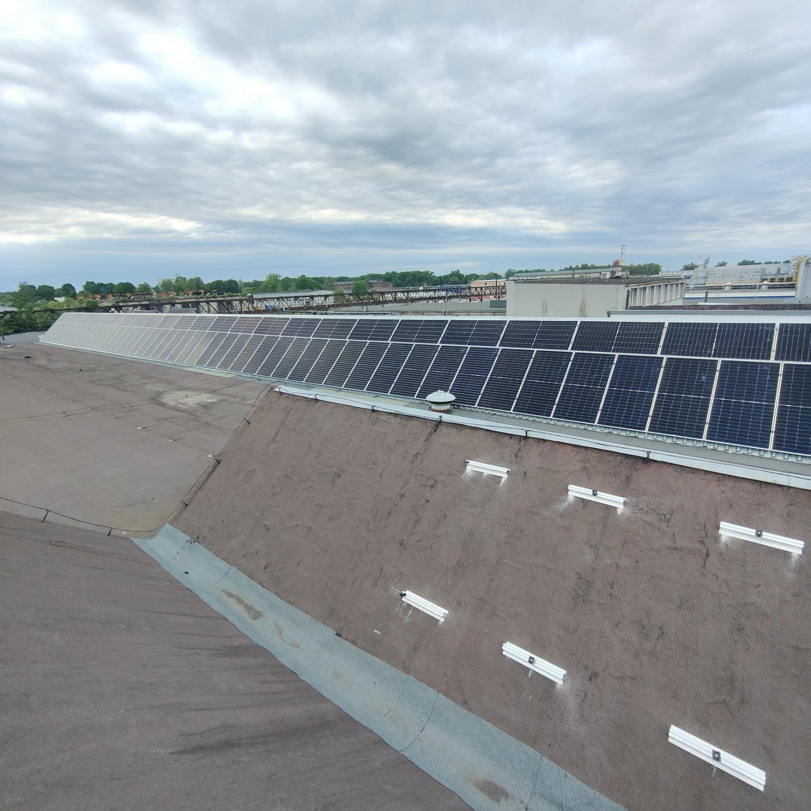 Rząd paneli słonecznych na płaskim dachu budynku przemysłowego, widok z góry na panele i system montażowy, pochmurne niebo w tle.