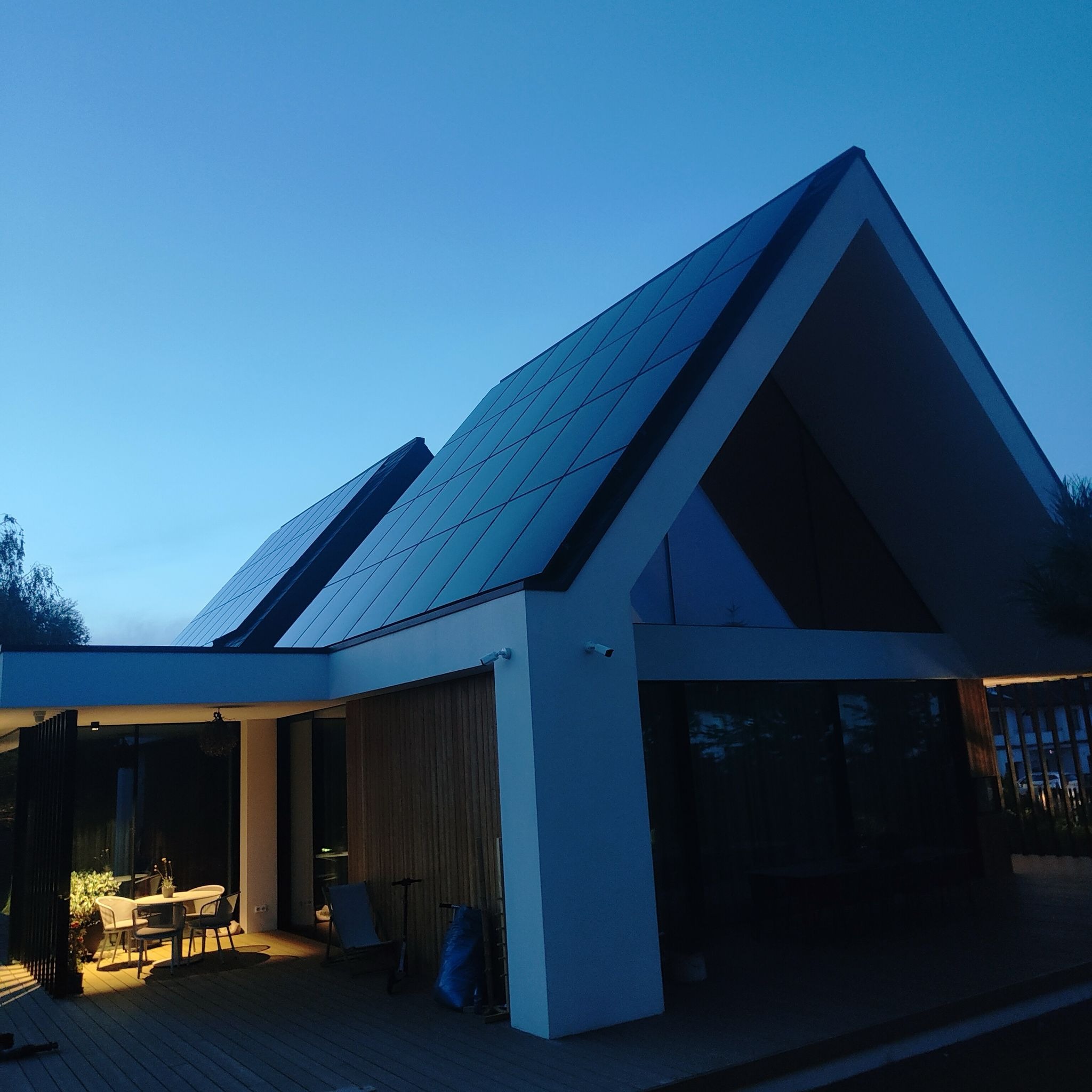 Nowoczesny dom jednorodzinny z panelami słonecznymi na dachu w wieczornym oświetleniu, widoczne kamery monitoringu na elewacji.