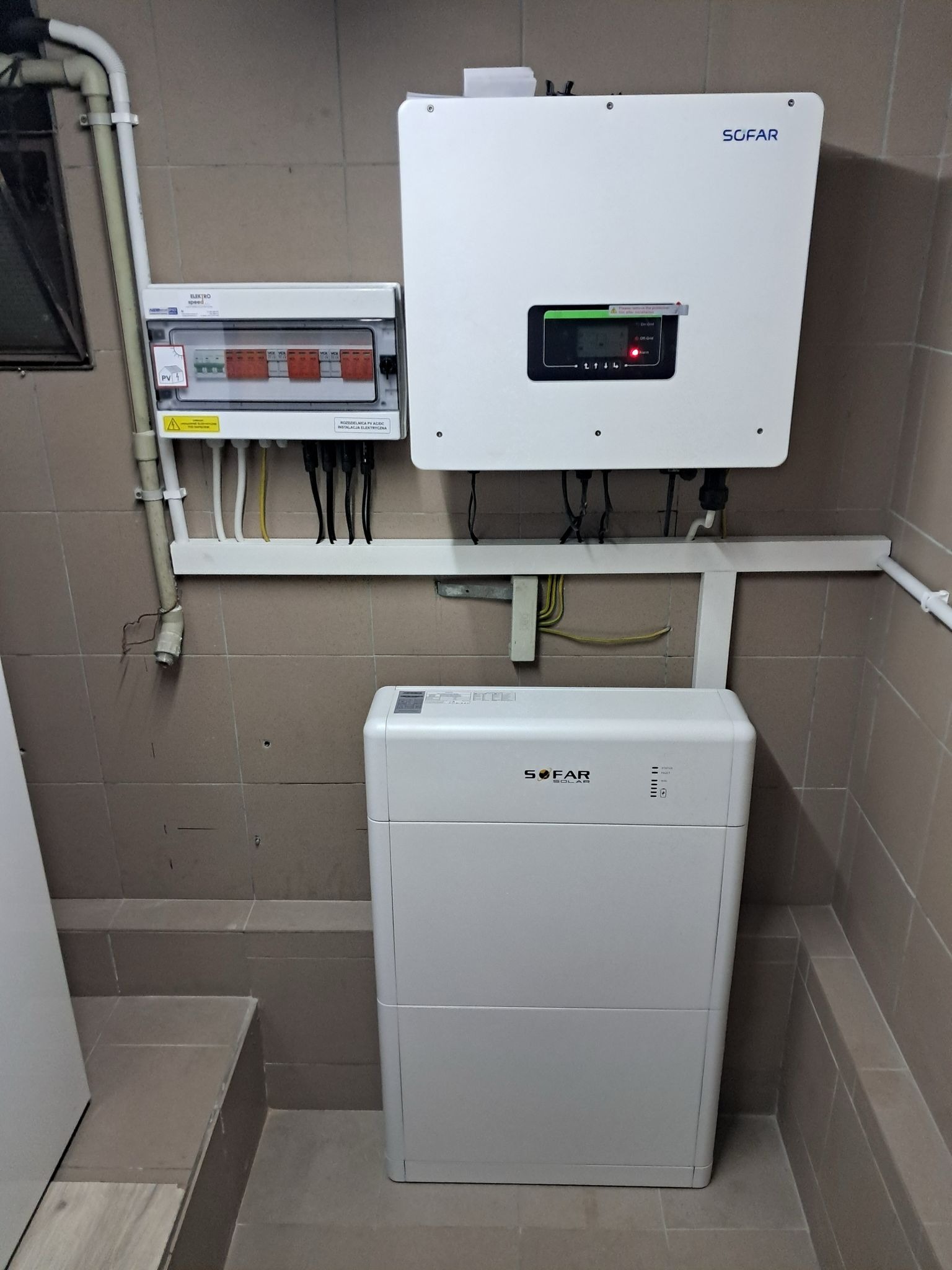 Biały inwerter i akumulator energii marki SOFAR zamontowane na kafelkowanej ścianie obok skrzynki elektrycznej z zabezpieczeniami.