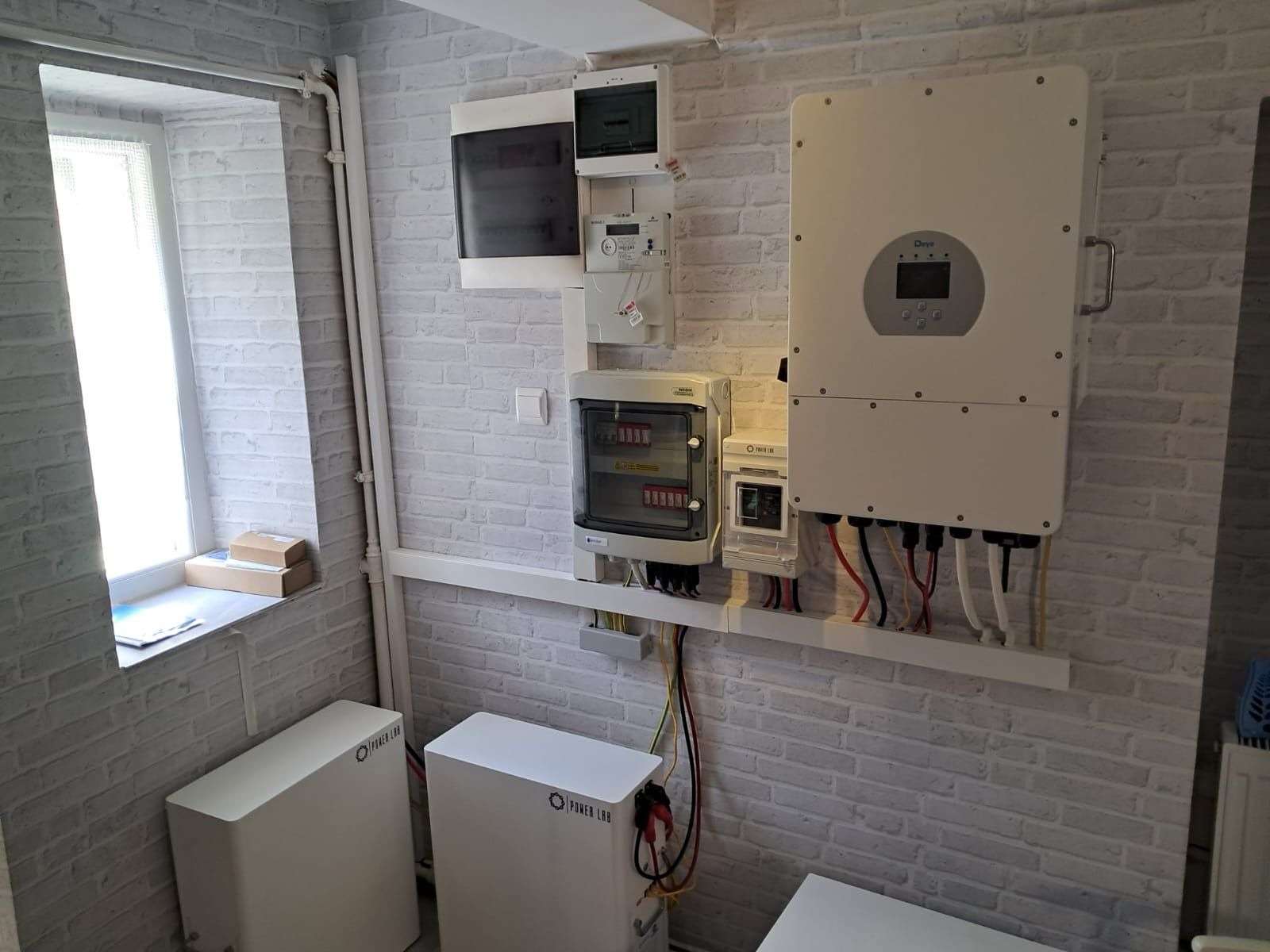 Instalacja wewnętrzna: białe magazyny energii Power Lab, inwerter Daye, rozdzielnice elektryczne i okablowanie na tle ściany z białej cegły w pomieszczeniu z oknem.