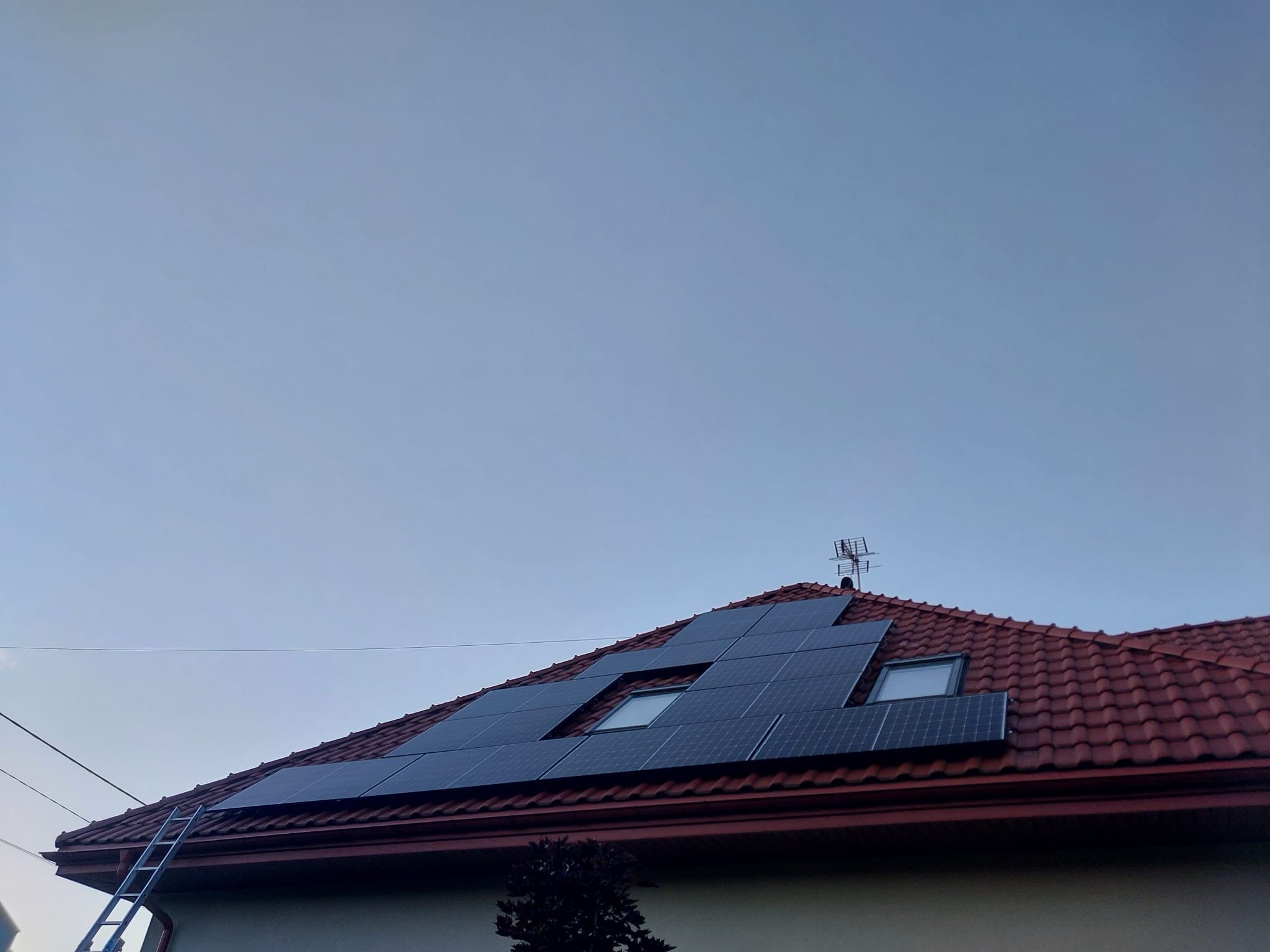 Panele fotowoltaiczne zamontowane na dachu z czerwonej dachówki, widoczna drabina oparta o rynnę, antena na dachu i fragment drzewa w dolnej części kadru.
