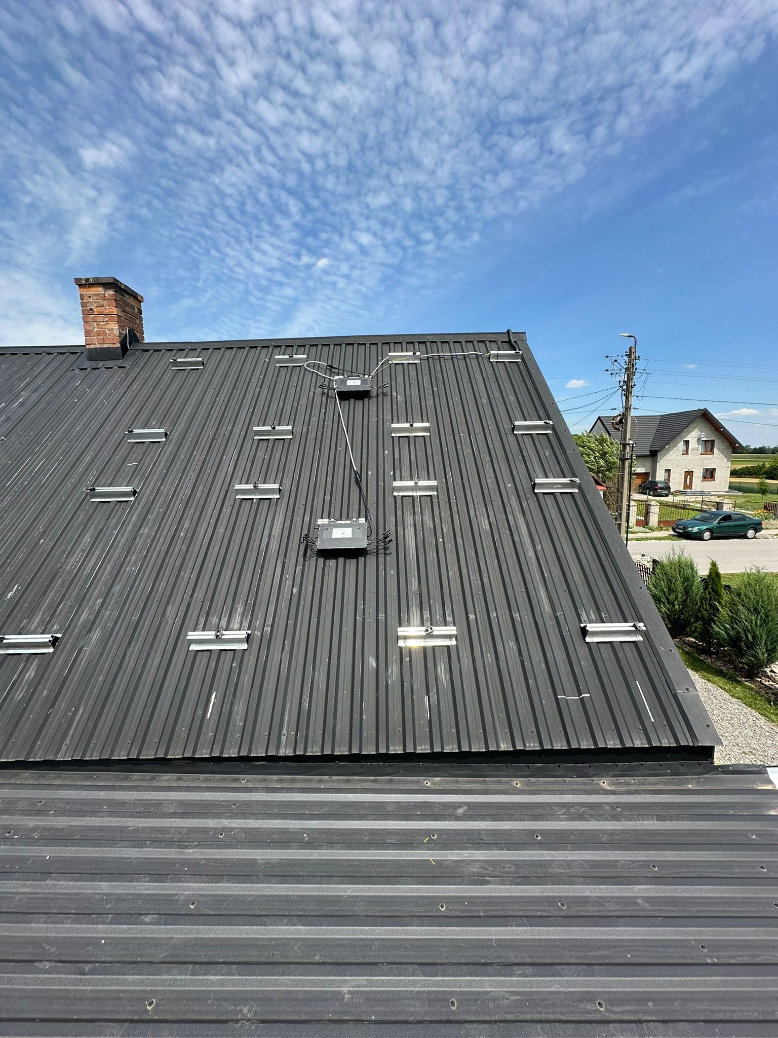 Czarny dach z blachy trapezowej z zamontowanymi szynami montażowymi i skrzynkami przyłączeniowymi, przygotowany do instalacji paneli słonecznych, widok z góry na tle błękitnego nieba i zabudowań.