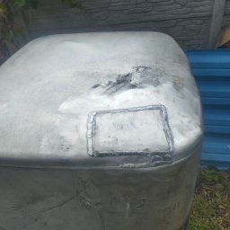 Jan-weld Usługi spawalnicze - Zbliżenie na zbiornik z metalu, z widocznymi śladami po niedawno zakończonym procesie łączenia elementów poprzez spawanie, w tle fragment niebieskiego płotu i zieleni.