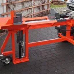 Pomarańczowa, mobilna łuparka do drewna z hydraulicznym siłownikiem, zaparkowana na kostce brukowej, z widocznym zaczepem przyczepy, kołami i fragmentem zaparkowanego samochodu w tle.