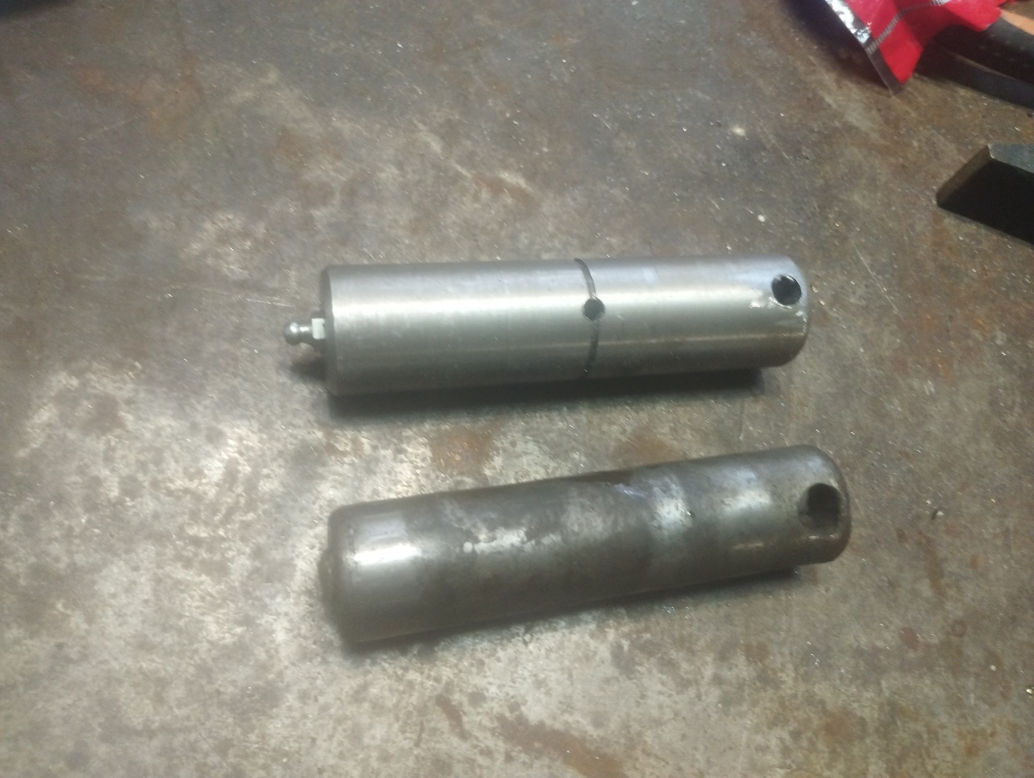 Dwa metalowe cylindryczne elementy z otworami, jeden nowy z kalamitką, drugi używany, leżące na metalowej powierzchni.