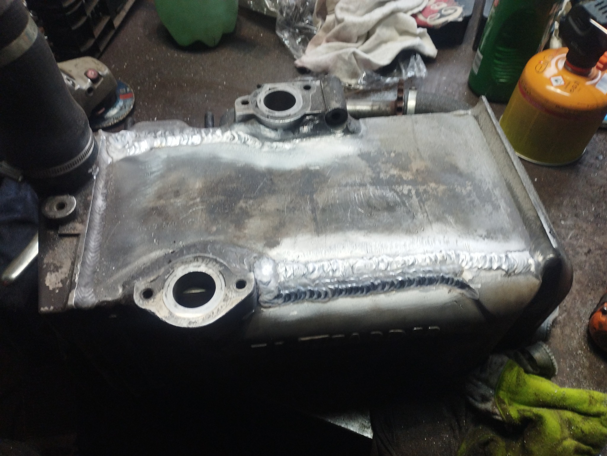 Metalowy zbiornik po naprawie, widoczne ślady zajarzenia łuku i spoiny, w tle narzędzia warsztatowe i butla z gazem.