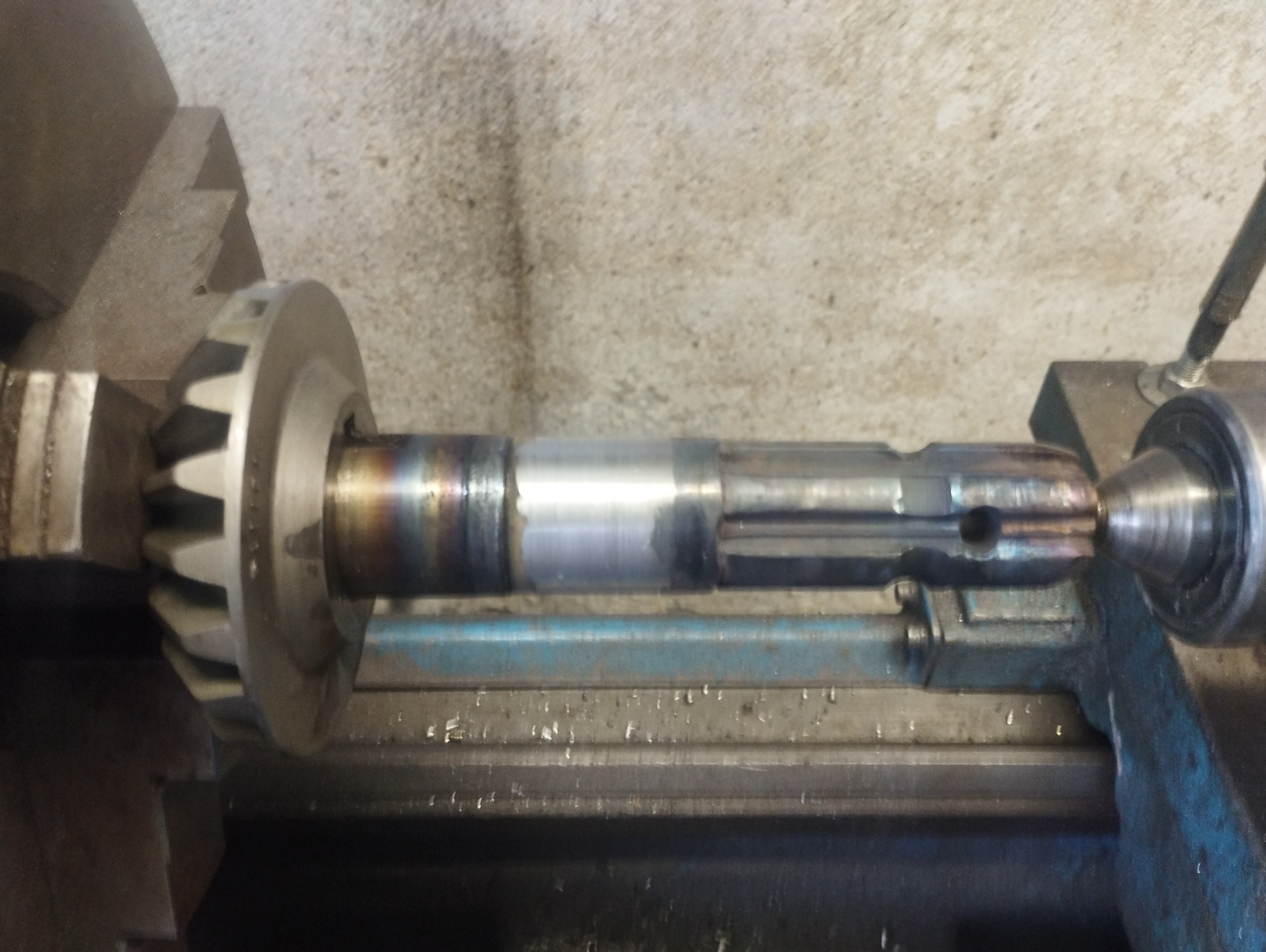Metalowy wał z zębatką zamocowany w tokarce, widoczne ślady obróbki i przypalenia na powierzchni.