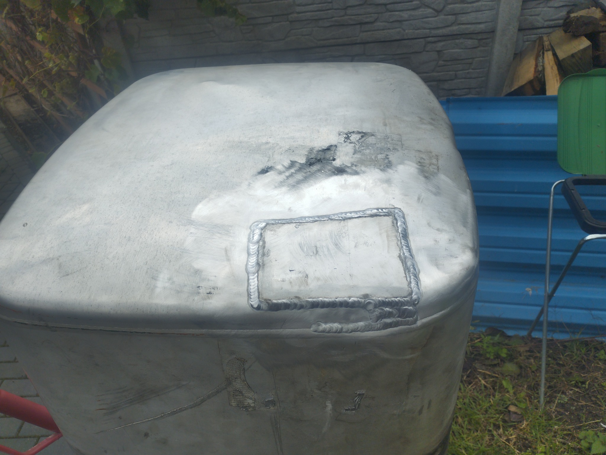 Zbliżenie na zbiornik z metalu, z widocznymi śladami po niedawno zakończonym procesie łączenia elementów poprzez spawanie, w tle fragment niebieskiego płotu i zieleni.