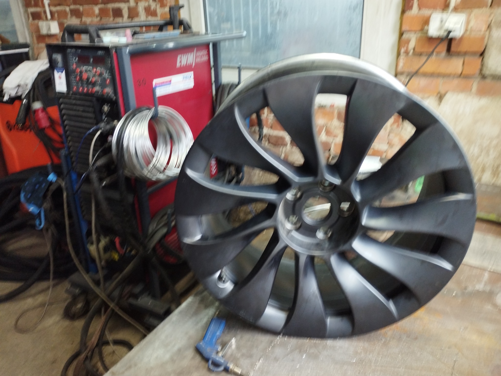 Szara felga aluminiowa z wieloma szprychami, leżąca na metalowym stole w warsztacie, z widocznym urządzeniem spawalniczym EWM i zwojem drutu spawalniczego w tle oraz pistoletem pneumatycznym obok.