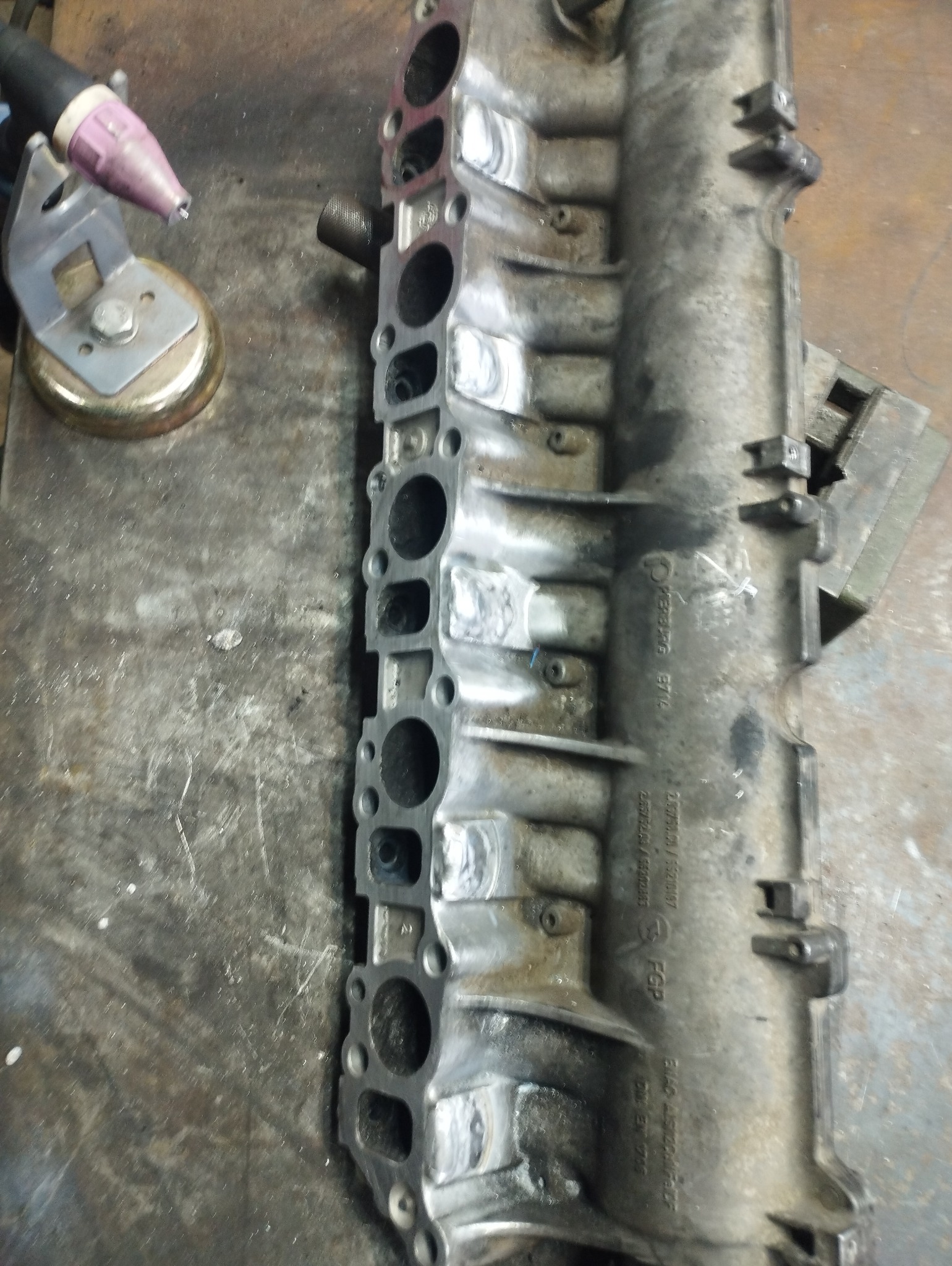 Spawany kolektor dolotowy silnika z widocznymi śladami spawania i uchwytem spawalniczym TIG obok, leżący na warsztatowym stole.