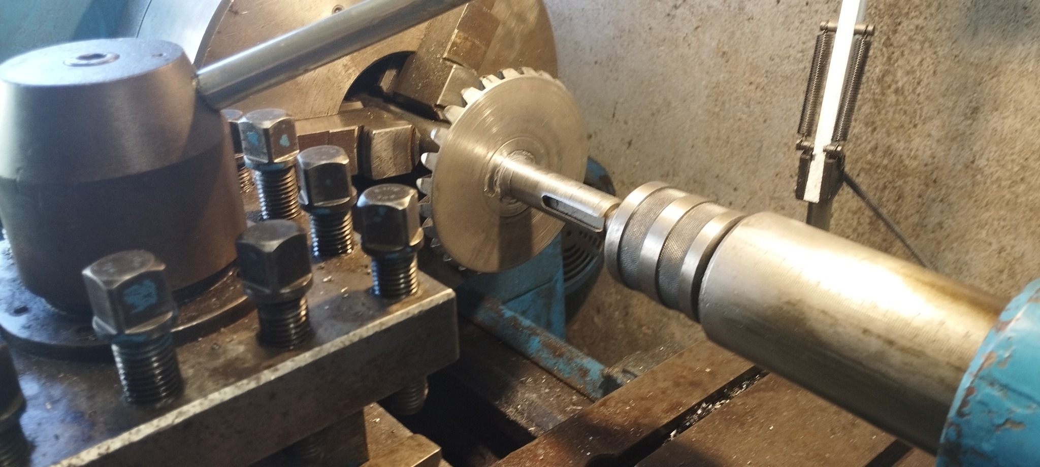 Metalowa zębatka zamocowana w uchwycie tokarskim podczas obróbki, widoczne ślady oleju i wióry na maszynie.