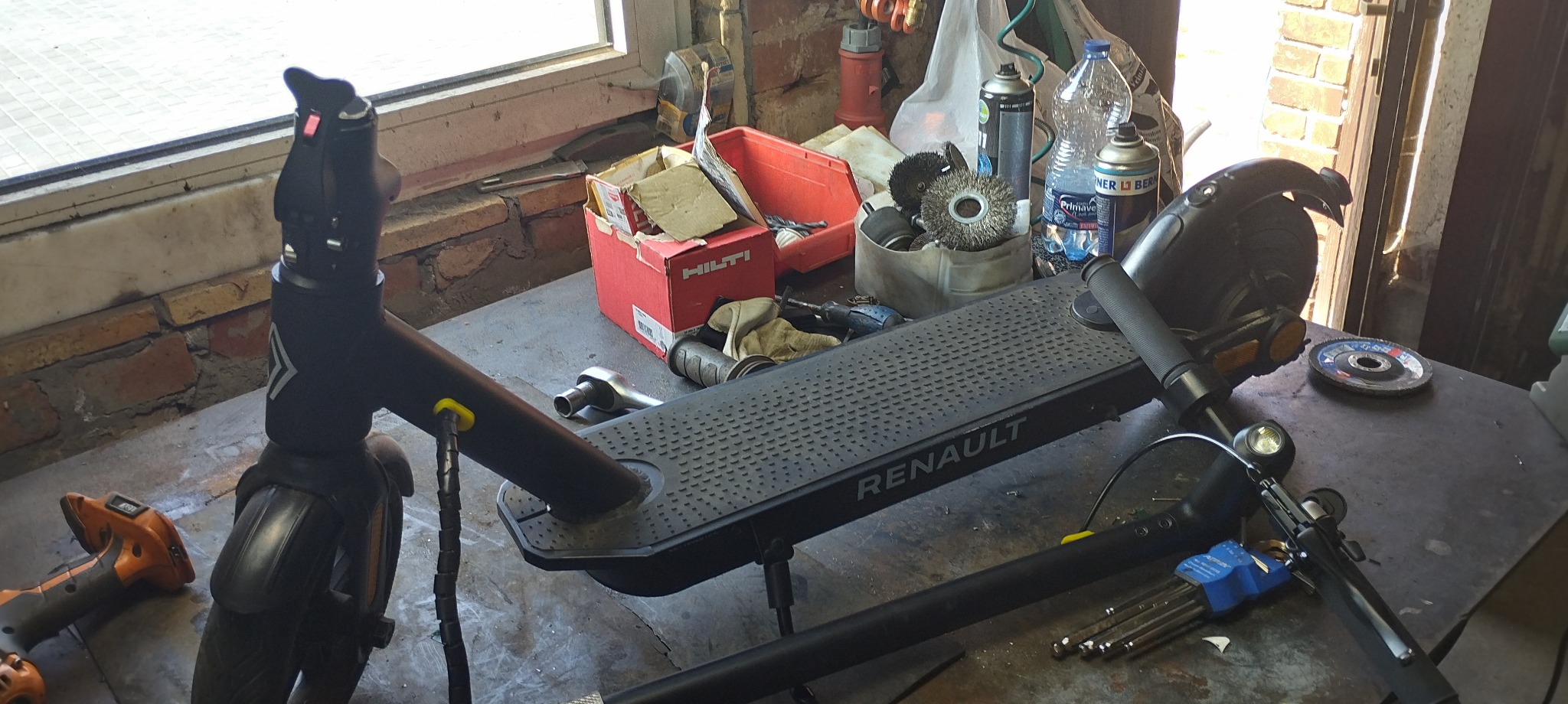 Elektryczna hulajnoga Renault na warsztatowym stole, otoczona narzędziami i akcesoriami, w tle ceglany mur i okno.