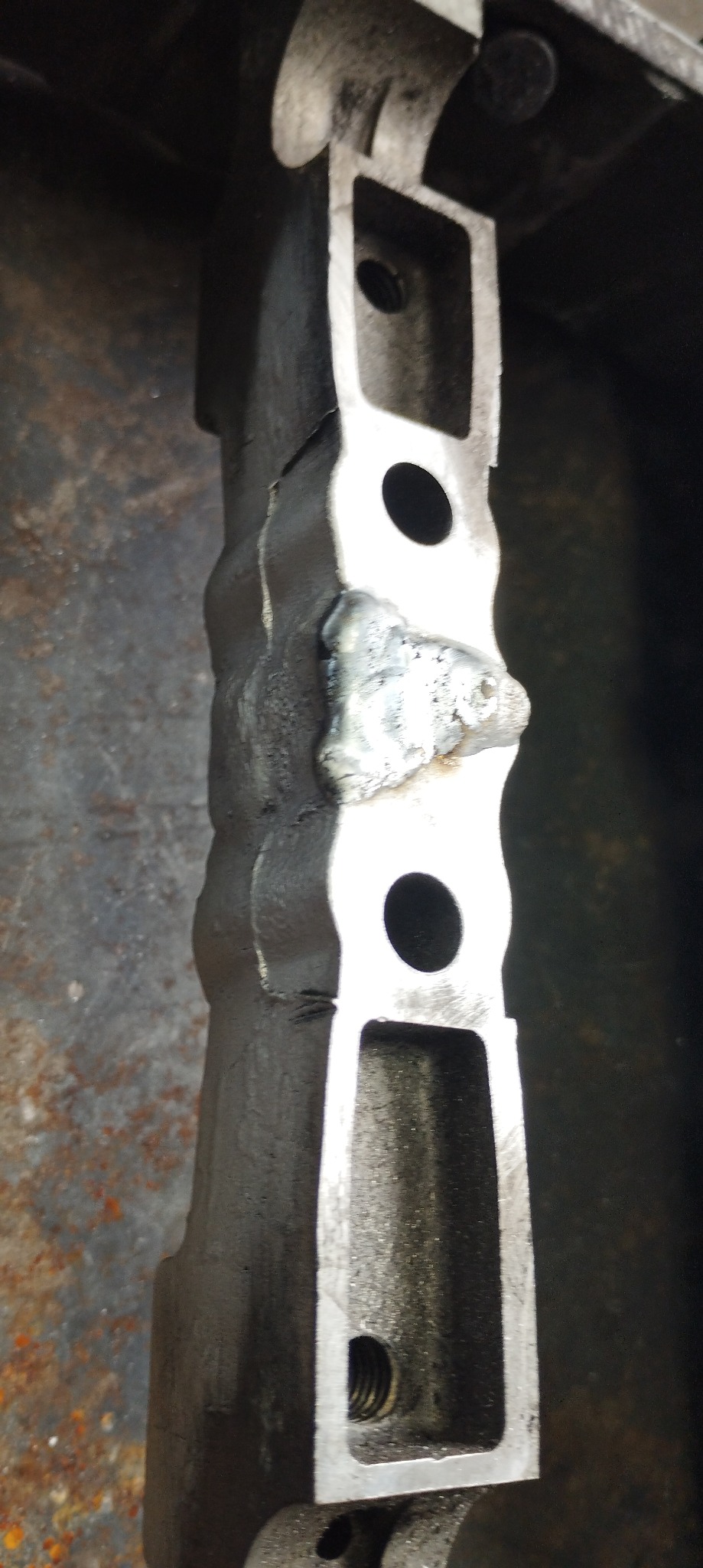 Szlifowany metalowy element konstrukcyjny z widocznym śladem po spawaniu oraz otworami, leżący na brudnym metalowym podłożu.