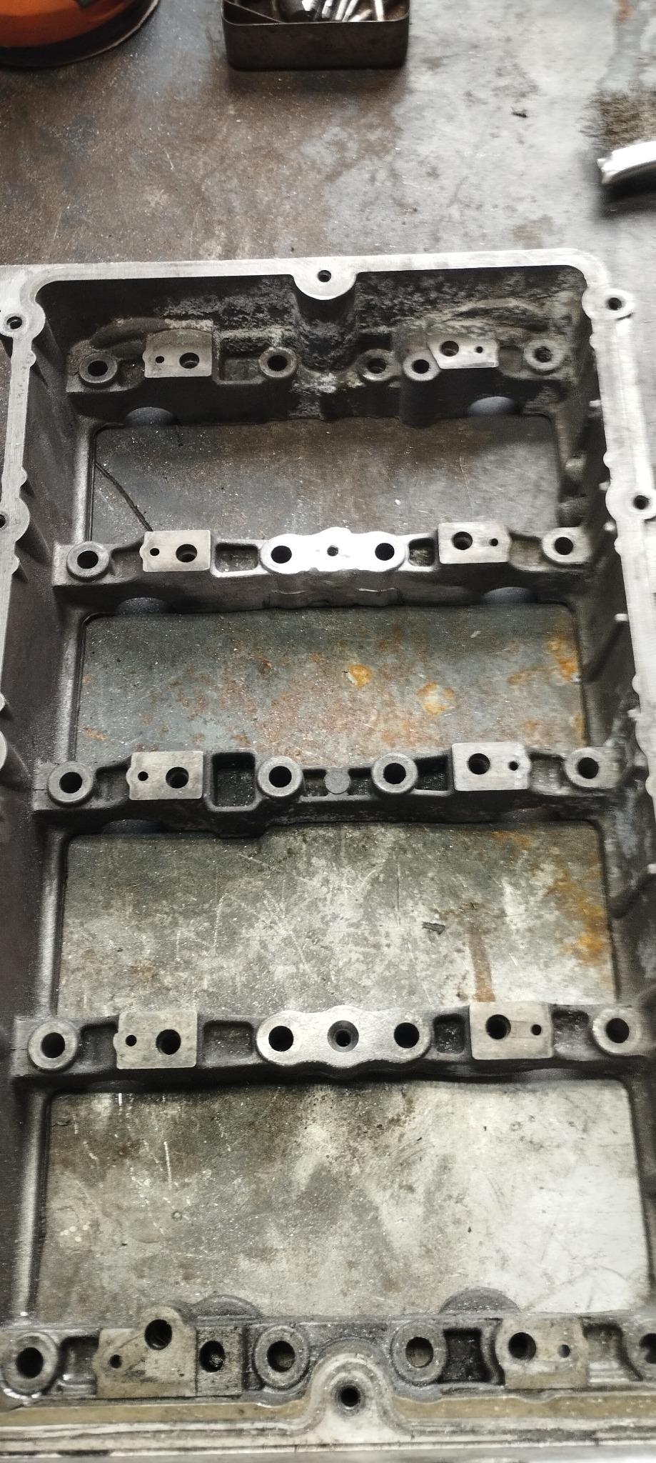 Szara, metalowa obudowa z licznymi otworami i śladami użytkowania, leżąca na brudnym warsztatowym stole.