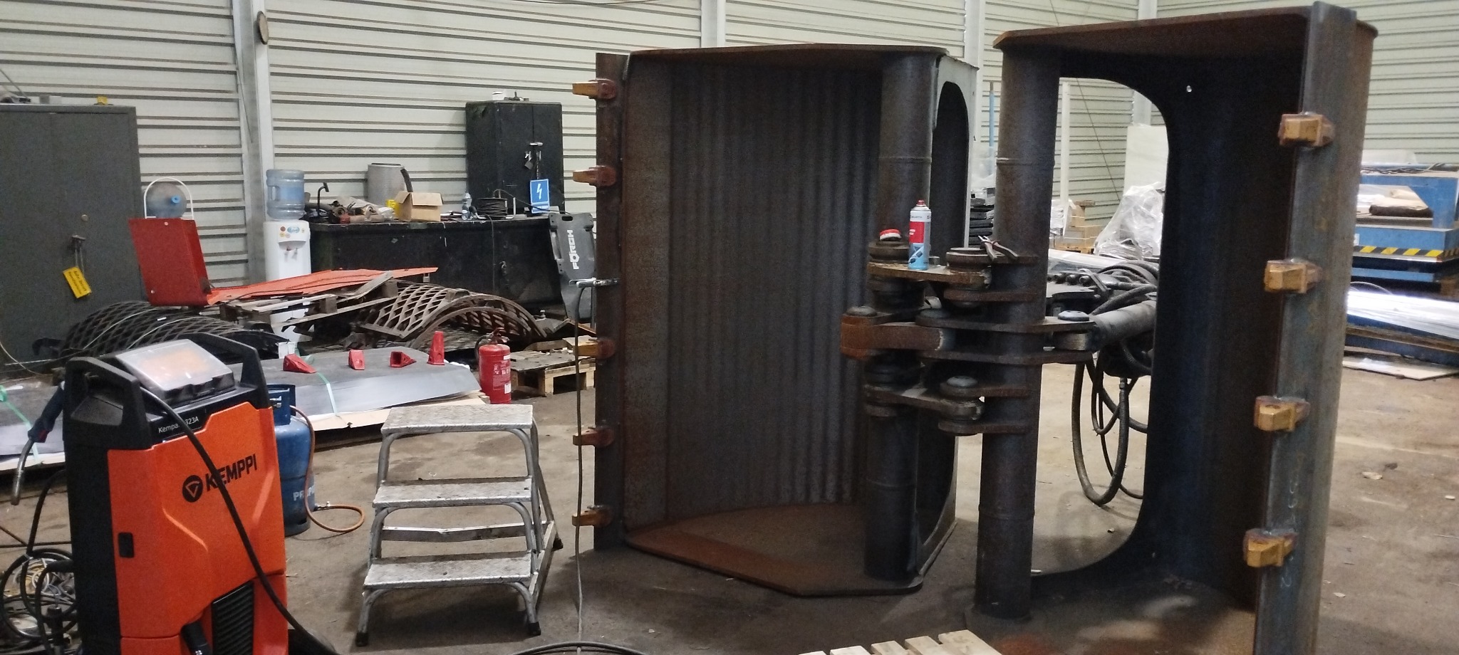 Dwie metalowe konstrukcje w warsztacie, z widocznymi spawami i elementami mocującymi, obok spawarka Kemppi i metalowe elementy do obróbki.