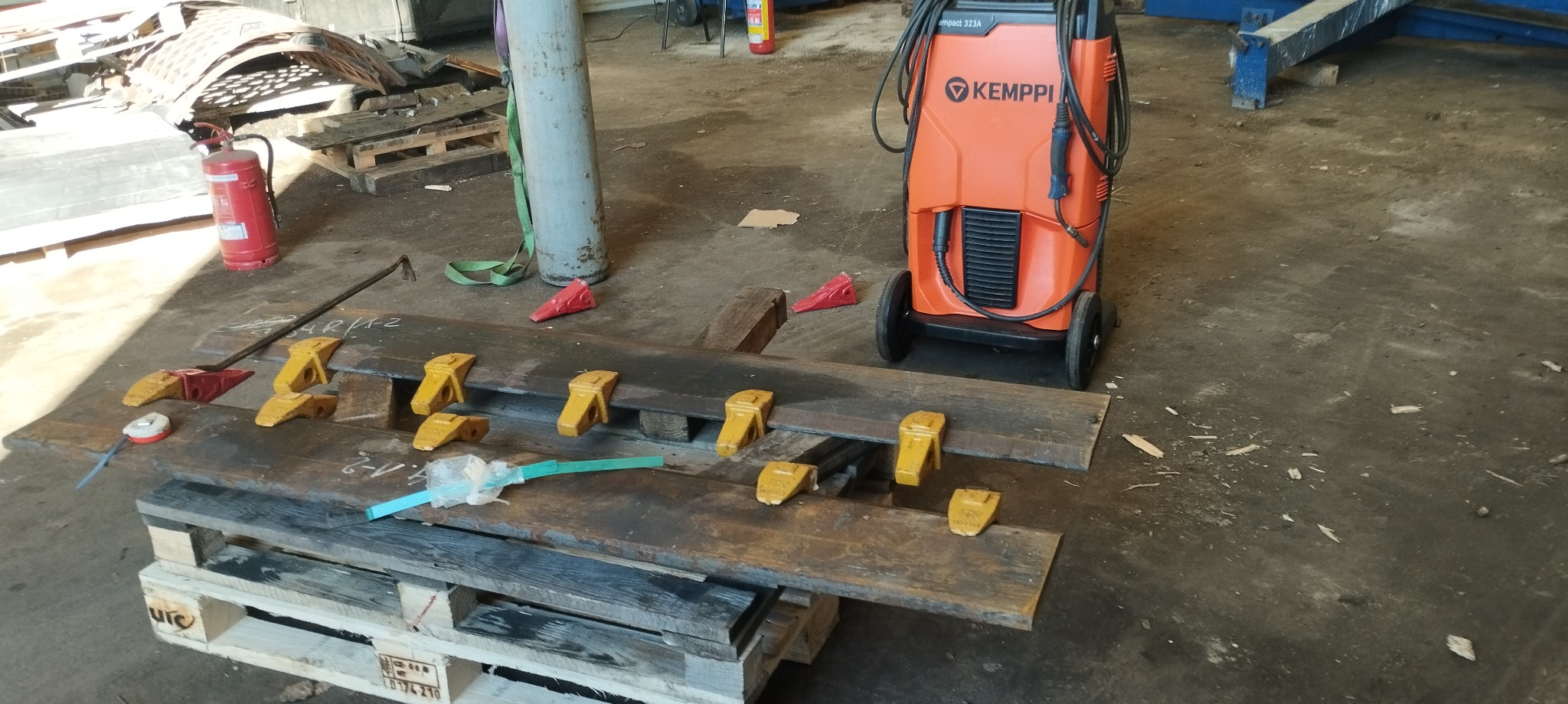 Przygotowane do obróbki metalowe elementy z żółtymi zębami mocującymi na drewnianych deskach, obok spawarka Kemppi, gaśnica i butla z gazem w warsztacie.