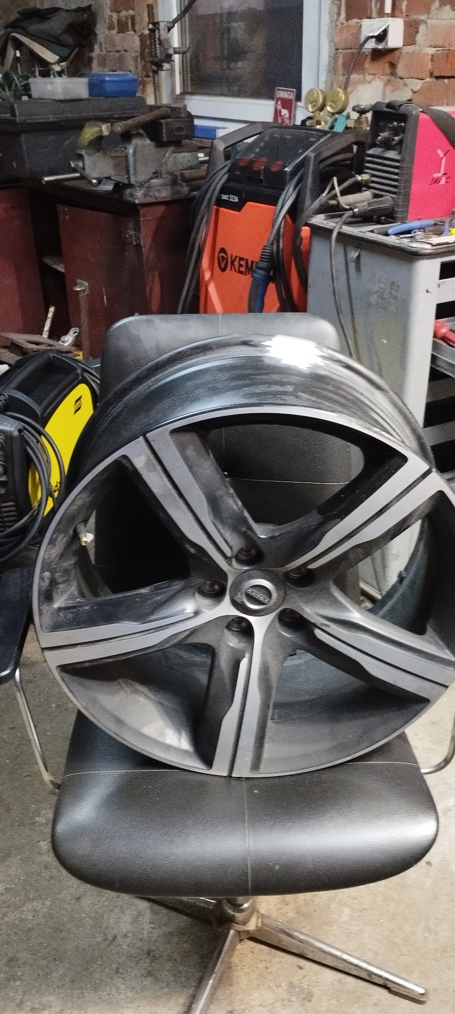Aluminiowa felga samochodowa Volvo z pięcioma ramionami, leżąca na czarnym krześle w warsztacie, widoczny sprzęt spawalniczy i narzędzia.