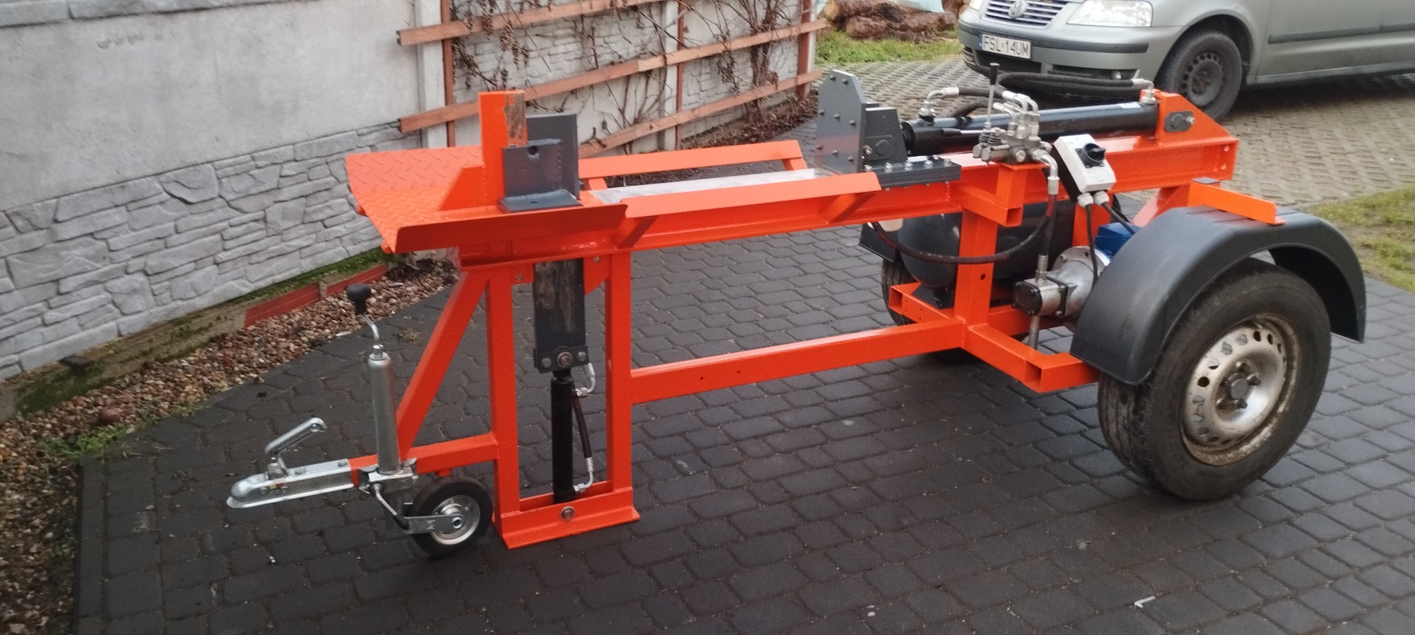 Pomarańczowa, mobilna łuparka do drewna z hydraulicznym siłownikiem, zaparkowana na kostce brukowej, z widocznym zaczepem przyczepy, kołami i fragmentem zaparkowanego samochodu w tle.