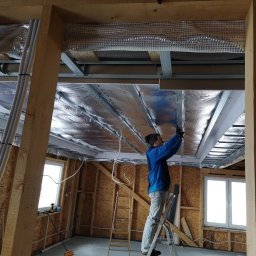 MULTI-FINISH - Pracownik na drabinie montuje folię termoizolacyjną na metalowym stelażu sufitu podwieszanego w pomieszczeniu z drewnianą konstrukcją ścian.