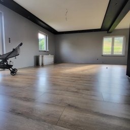 MULTI-FINISH - Pokój po remoncie z panelami podłogowymi w odcieniach brązu, szare ściany, czarny sufit podwieszany i wózek dziecięcy w rogu.