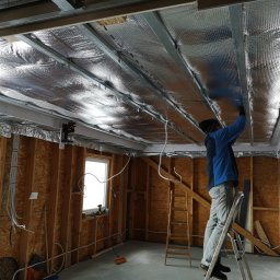 MULTI-FINISH - Prace wykończeniowe: montaż izolacji termicznej pod sufitem z użyciem folii aluminiowej i metalowych profili, widoczny pracownik na drabinie i surowe ściany z instalacjami.