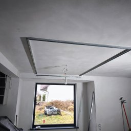 MULTI-FINISH - Surowy sufit podwieszany ze stelażem z profili metalowych, widok z dołu, w tle okno z widokiem na podwórko, w pomieszczeniu widoczne narzędzia i materiały budowlane.