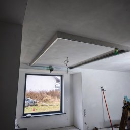 MULTI-FINISH - Wnętrze w trakcie prac wykończeniowych: sufit podwieszany w trakcie montażu, widoczne profile i płyty, ściany przygotowane pod malowanie, drabina z narzędziami, okno z widokiem na zewnątrz.