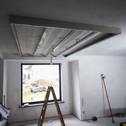 MULTI-FINISH - Montaż stelaża sufitu podwieszanego w surowym, jasnym pomieszczeniu z oknem; widoczna drabina, profile metalowe, płyty gipsowe i podpory teleskopowe.