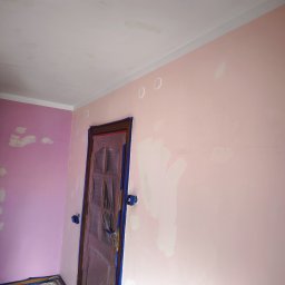 MULTI-FINISH - Różowe ściany w trakcie przygotowania do malowania: zagruntowane ubytki, zabezpieczone drzwi folią i taśmą malarską, widoczne ślady po szpachlowaniu na suficie.