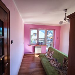 MULTI-FINISH - Pokój po malowaniu na różowo, widoczny fragment sofy z kocem w koty, okno z parapetem i grzejnikiem, drewniana podłoga i drzwi.