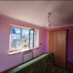 MULTI-FINISH - Pokój z fioletowymi ścianami, otwartym oknem z widokiem na zewnątrz, grzejnikiem pod oknem i zieloną kanapą z kocem w koci wzór.