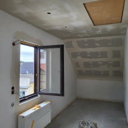 MULTI-FINISH - Pokój na poddaszu w trakcie prac wykończeniowych: ściany i sufit po szpachlowaniu, widoczne łączenia płyt gipsowo-kartonowych, okno otwarte na zewnątrz, zabezpieczony grzejnik pod oknem.