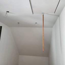 MULTI-FINISH - Białe wnętrze poddasza w trakcie malowania, widoczne otwory na oświetlenie, klapa strychowa z haczykiem i wiszącą listwą, surowe ściany.