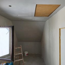 MULTI-FINISH - Poddasze w trakcie prac wykończeniowych: ściany i sufit po szpachlowaniu, widoczna drewniana klapa włazu strychowego, drabina oparta o okno.