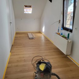 MULTI-FINISH - Pokój na poddaszu w trakcie układania paneli podłogowych, ściany zabezpieczone taśmą malarską, widoczny odkurzacz przemysłowy i paczka paneli.