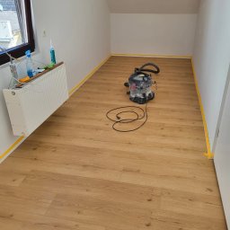 MULTI-FINISH - Pokój na poddaszu z nową, jasną podłogą z paneli, zabezpieczony taśmą malarską przy ścianach, z odkurzaczem przemysłowym na środku i grzejnikiem z akcesoriami malarskimi w rogu.