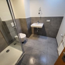 MULTI-FINISH - Nowoczesna łazienka z szarymi płytkami, kabiną prysznicową ze srebrnymi ramami, wiszącą umywalką i toaletą w stylu minimalistycznym oraz dekoracyjną lampą wiszącą z żarówką na kablu.