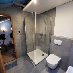 MULTI-FINISH - Nowoczesna łazienka z kabiną prysznicową ze szklanymi ściankami, wiszącą toaletą i szarą płytką na ścianach, widoczne ślady prac wykończeniowych przy futrynie drzwi.